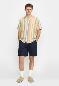 Korte mouwen overhemd met verticale strepen in pastelkleuren, met knoopsluiting aan de voorkant, gecombineerd met marineblauwe shorts en beige instap schoenen. Witte sokken zichtbaar.
