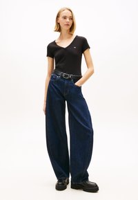 Melna T-krekls ar V-veida izgriezumu, augsta vidukļa vaļīgas tumši zilas džinsa bikses, melns jostas siksna ar sudraba sprādzi, apjomīgas melnas kurpes, minimālistisks fons.