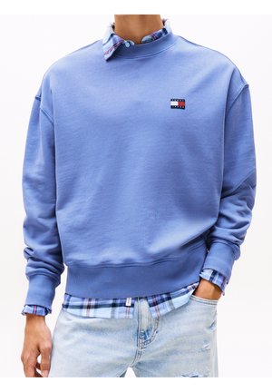 Mand iført en lyseblå Tommy Jeans sweatshirt over en blå ternet skjorte med lyseblå revnede jeans, hånden i lommen.