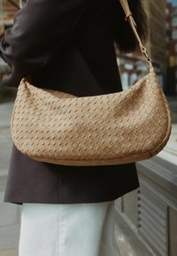 Geweven beige handtas met een ronde vorm, voorzien van een gladde textuur en een slanke schouderband die wordt vastgezet met een metalen sluiting.