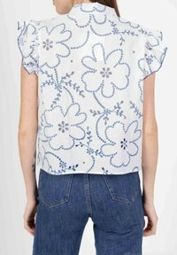 Blouse blanche à manches courtes avec broderie florale bleue et motifs circulaires, présentant des manches volantées et un col rond. Vue de dos visible.