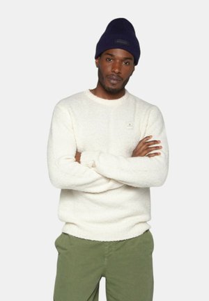 Un pull moelleux de couleur crème avec un col rond et des poignets côtelés, porté avec un pantalon vert olive. Un petit logo est visible sur la poitrine.