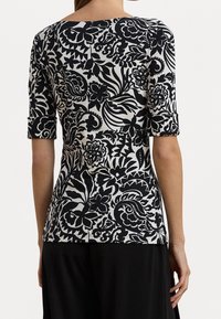 Woman wearing a black and white floral patterned shirt with elbow-length sleeves and black pants, viewed from the back against a white background.