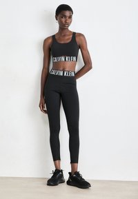 Soutien-gorge de sport noir avec un bandeau 'CALVIN KLEIN', associé à un legging noir taille haute avec un logo similaire. Le modèle porte des baskets noires.