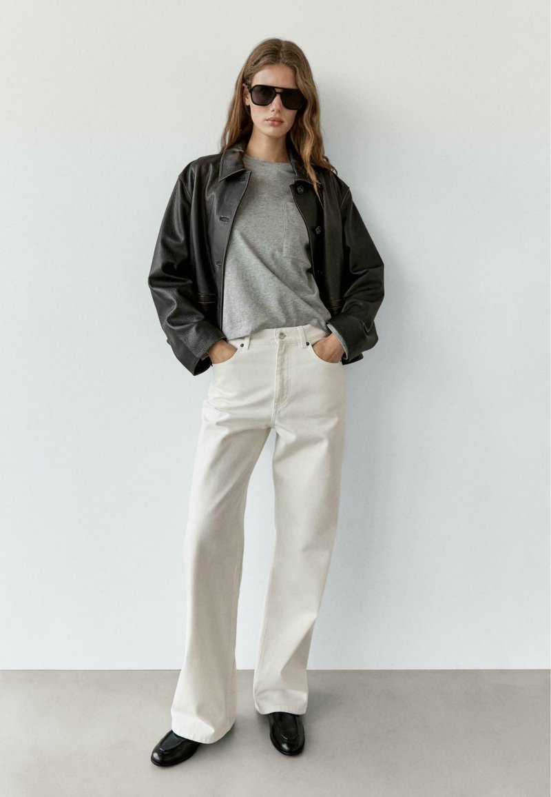 Svart läderjacka, grå t-shirt, vita jeans med vida ben, svarta skor och stora solglasögon; minimalistisk design, släta texturer, neutrala färger.