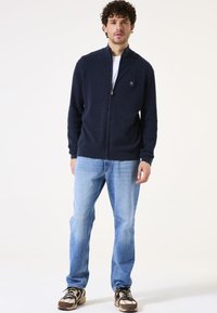 Chaqueta de punto con cremallera de color navy, con superficie texturizada, cuello alzado y un pequeño logo en el pecho, combinada con jeans azul claro y zapatos estampados.