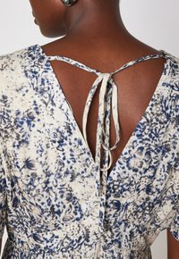 Abito floreale in crema con accenti blu e grigi. Presenta un design con scollo a V sul retro con sottili laccetti, realizzato in tessuto leggero.