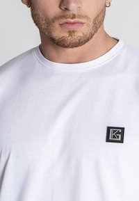 Weißes T-Shirt mit kurzen Ärmeln aus Baumwolle, mit einem schwarzen quadratischen Patch und grauem Logo auf der linken Brustseite. Glatte Textur, Rundhalsausschnitt.