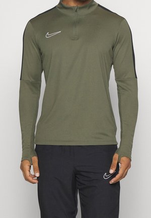 Homme portant un haut de sport à manches longues vert olive avec une bande noire sur les manches et un pantalon noir, tous deux ornés de logos Nike blancs.