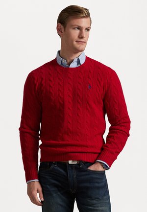 Roter Kabelstrickpullover mit gerippten Bündchen und Saum, ausgestattet mit einem kleinen gestickten Logo auf der Brust. Über einem hellblauen Hemd mit Kragen getragen.