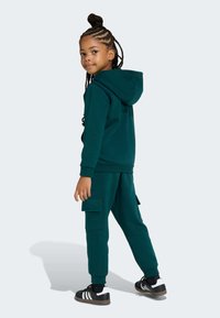 Ensemble sweat-shirt et pantalon vert en tissu doux, avec une capuche, des poignets côtelés et des poches cargo. Des baskets noires et blanches complètent le look.