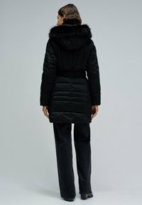 Chaqueta negra acolchada con capucha de piel sintética, cintura ajustada, textura acolchada y mangas largas. La modelo lleva pantalones anchos azul oscuro.