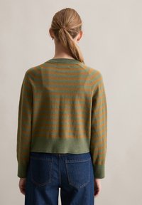 Vrouw met blond haar in een lage paardenstaart, gekleed in een groenbruin gestreepte trui en donkerblauwe spijkerbroek, met haar rug naar de camera tegen een effen achtergrond.