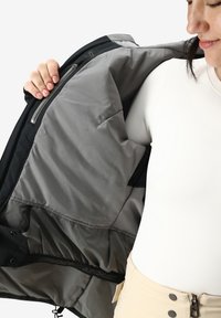 L'interno della giacca nera presenta una fodera grigia liscia, una tasca con zip e dettagli elastici. L'outfit del modello include una maglietta aderente bianca e pantaloni beige.