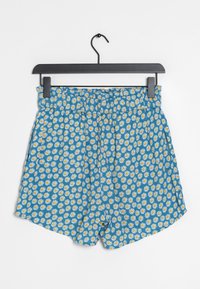 Shorts bleus à motif floral de marguerites blanches et jaunes, taille élastique, coupe ample et texture lisse. Suspendus à un crochet noir.