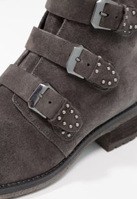 Bottine en daim gris avec trois brides à boucle métallique, détails cloutés et un talon bas empilé.