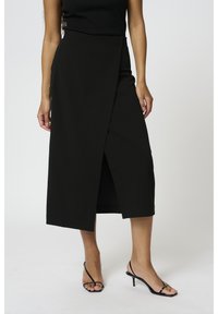 Jupe noire enveloppante à taille haute, coupe mi-genou, texture douce et fente à l'avant, associée à des sandales à talons hauts noires à lanières.