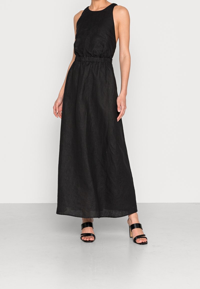 Femme portant une robe maxi noire sans manches avec une taille cintrée et des sandales à talons noires à brides, debout sur un sol gris.