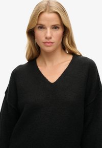 Suéter negro de cuello en V, hecho de una tela suave y texturizada con mangas de corte holgado. El diseño presenta una silueta relajada y un estilo minimalista.