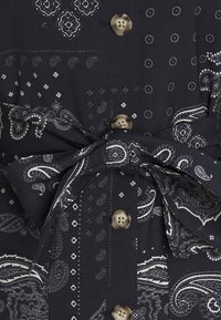 Tmavá tkanina s bílými vzory paisley a geometrickými vzory, béžové knoflíky uprostřed a uvázaný pás z tkaniny v pase.