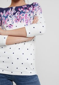 Witte lange mouwen top met een bloemenpatroon in roze en blauw op de halslijn en marineblauwe stippen op het lichaam. Zacht materiaal met een klassieke pasvorm.