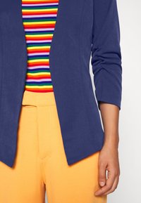 Blazer bleu marine aux manches retroussées porté sur un haut rayé multicolore et un pantalon taille haute orange vif, présentant une texture de tissu lisse.