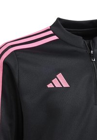 Černá zipová bunda s růžovými pruhy podél ramen a vyšívaným růžovým logem. Materiál vypadá hladce, s pevným límcem.