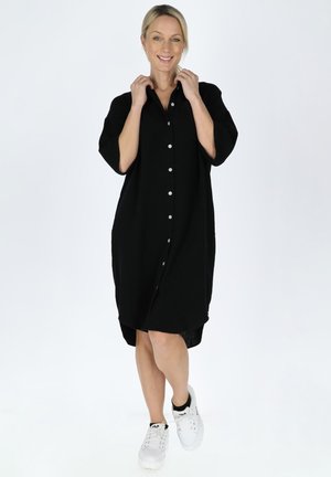 Robe boutonnée noire à manches courtes, avec une coupe ample et un ourlet asymétrique plus long à l'arrière. Portée avec des baskets blanches. Texture lisse.