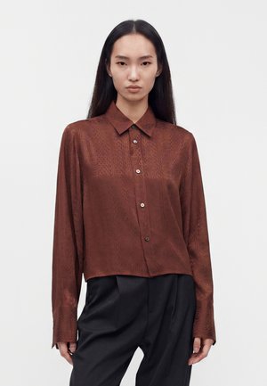 Femme aux longs cheveux noirs portant une chemise marron texturée boutonnée et un pantalon noir, debout devant un fond blanc uni.