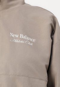 Svijetlosmeđa jakna na vjetar sa zip ovratnikom i izvezenim logotipom "New Balance Athletic Club" u bijeloj boji. S mekanom teksturom i minimalističkim dizajnom.