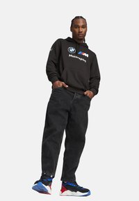 Black hoodie with BMW Motorsport logo, paired with loose black jeans and multi-colored sneakers featuring blue, red, and white accents.