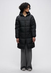 Crna puhasta jakna s kapuljačom, s patentnim zatvaračem sprijeda, širokog kroja, prošivenim uzorkom i dugim krojem koji seže do sredine lista.