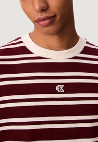 Svetr se pruhy v barvách maroon a krémová, s žebrovaným kulatým výstřihem a malým vyšitým logem na prsou. Měkká struktura, slim fit design.