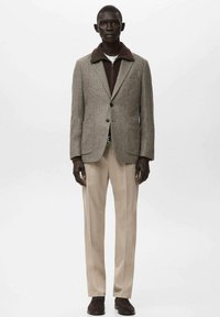 Hnědý tweedový blejzr s kontrastním tmavým límcem, doplněný o tmavý pletený svetr, krémové kalhoty a hnědé boty. Jednobarevné pozadí.