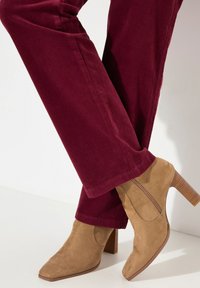 Stivaletti alla caviglia in suede beige con punta appuntita e tacco a blocco rigato marrone, abbinati a pantaloni in corda bordeaux. Dettaglio con zip sugli stivaletti.