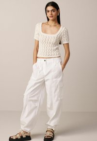 Haut tricoté couleur crème à manches courtes avec un décolleté carré ; associé à un pantalon cargo blanc ample avec poches et des sandales noires.