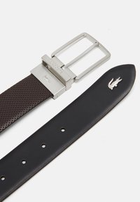Brun läderbälte med strukturerade perforeringar. Silvermetallspänne med graverad "Lacoste." Svart läderrem som har en silverkrokodil-logotyp.