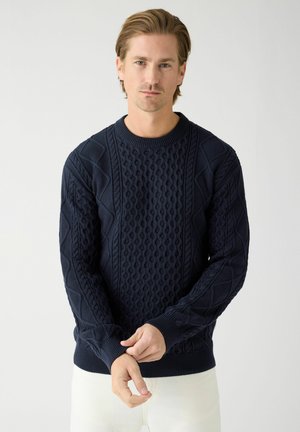 Mand justerer manchet på mørkeblå kabelstrikket sweater, stående mod ensfarvet lyst gråt baggrund.