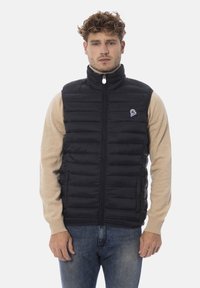 Schwarze Steppweste mit vertikaler Steppung, hohem Kragen, durchgehendem Reißverschluss und kleinem Logopatch auf der linken Seite. Über einem beigen Hemd getragen.