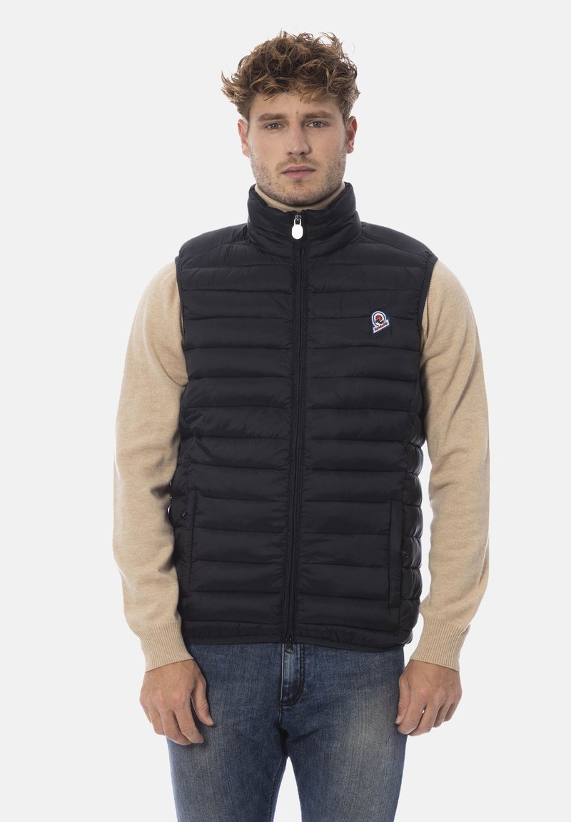 Schwarze Steppweste mit vertikaler Steppung, hohem Kragen, durchgehendem Reißverschluss und kleinem Logopatch auf der linken Seite. Über einem beigen Hemd getragen.