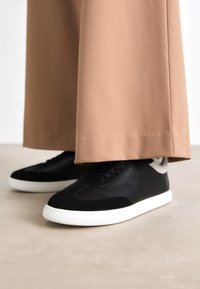 Svarte og grå joggesko med en glatt læroverdel og myke semskede detaljer; hvit gummisåle; brukt med beige vide bukser.