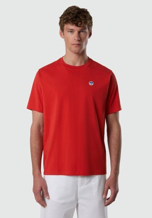 Joven con cabello corto y rizado que lleva una camiseta roja de manga corta lisa con un pequeño logo redondo azul y blanco en el pecho.