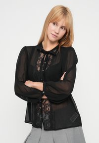 Zwarte doorschijnende blouse met kanten inzetstukken, knopen voor, striksluiting bij de kraag en pofmouwen. Gestructureerde stof met fijne details.