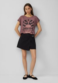 Grafisches T-Shirt in gedämpftem Rosa mit dem Text "SMILE CLUB", kombiniert mit einem schwarzen Wickelrock. Flache schwarze Schuhe runden das Outfit ab.