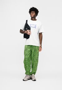 Fehér póló lila grafikával, zöld mintás nadrág és szürke tornacipő. A modell egy fekete táskát tart. Simogató textúrák és laza szabás.