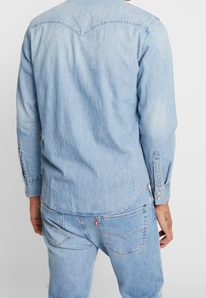 Mann trägt hellblaues Jeanshemd und Jeans, von hinten gezeigt mit sichtbaren Nähten und Knöpfen an den Hemdmanschetten.