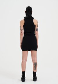 Schwarzes, geripptes, ärmelloses Kleid mit taillierter Passform, das bis zur Mitte des Oberschenkels reicht, getragen mit schwarzen, knielangen Socken und Plateauschuhen; sichtbare Tattoos an den Beinen.