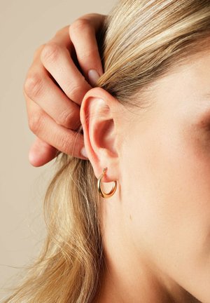 Nahaufnahme eines Ohrs mit einem goldenen Creolen-Ohrring, Hand zieht blondes Haar zurück vor einem beigen Hintergrund.