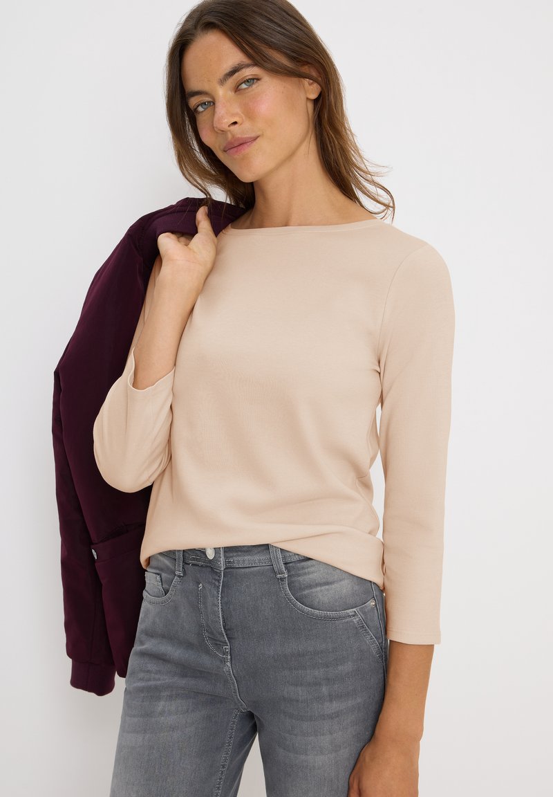 Haut beige à manches longues avec un col rond, associé à un jeans gris. Une veste bordeaux foncé est drapée sur l'épaule.