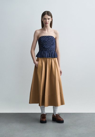 Νεαρή γυναίκα στέκεται με τα χέρια στις τσέπες, φορώντας μπλε τοπ με πουά χωρίς τιράντες, μπεζ φούστα μέτριου μήκους, λευκές κάλτσες και καφέ παπούτσια πλατφόρμα.